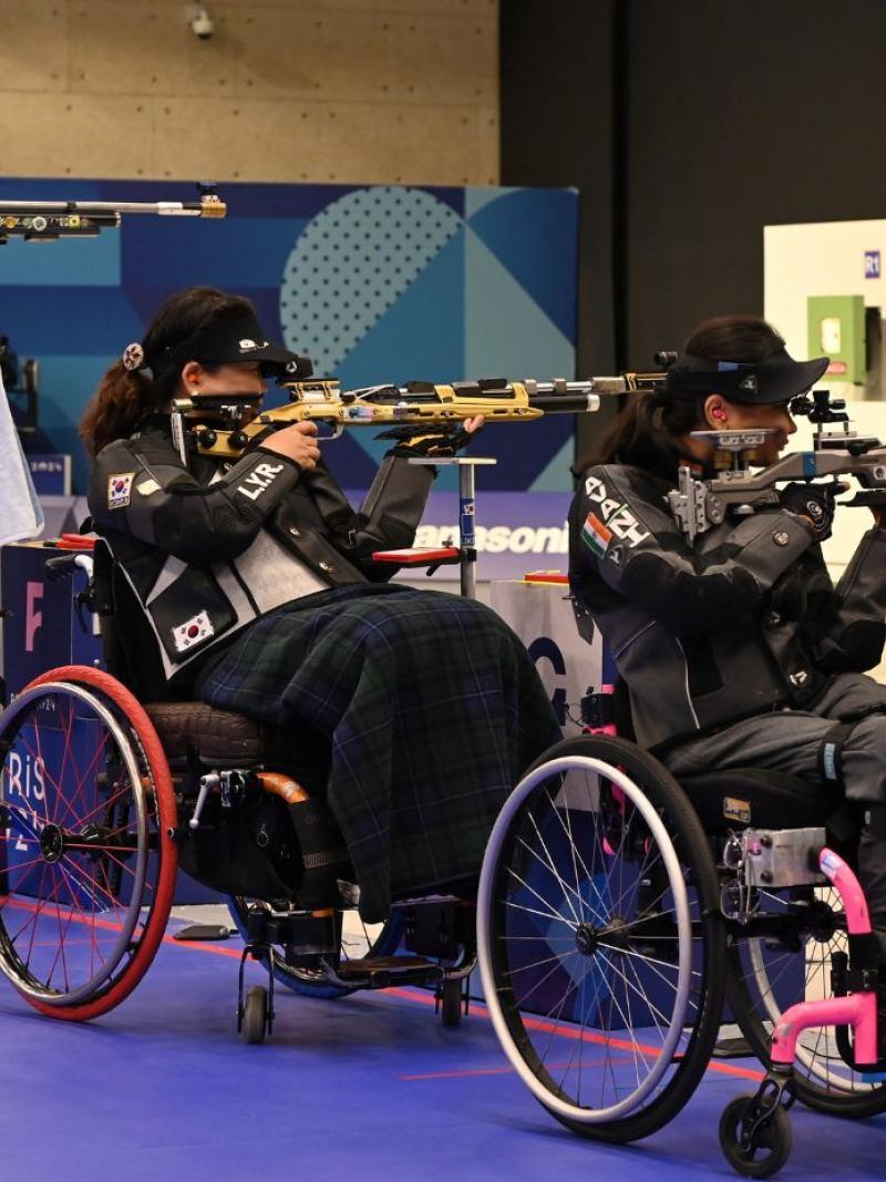 A standing woman and two women in wheelchairs competing in a rifle shooting range